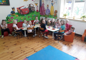 Zdjęcie przedstawia dzieci siedzące przy stolikach na czerwonych kanapach. Na stoliku, w plastikowych talerzykach znajdują się kolorowe pisanki. Za dziećmi na ścianie widać namalowane obrazki przedstawiające postacie bajkowe.